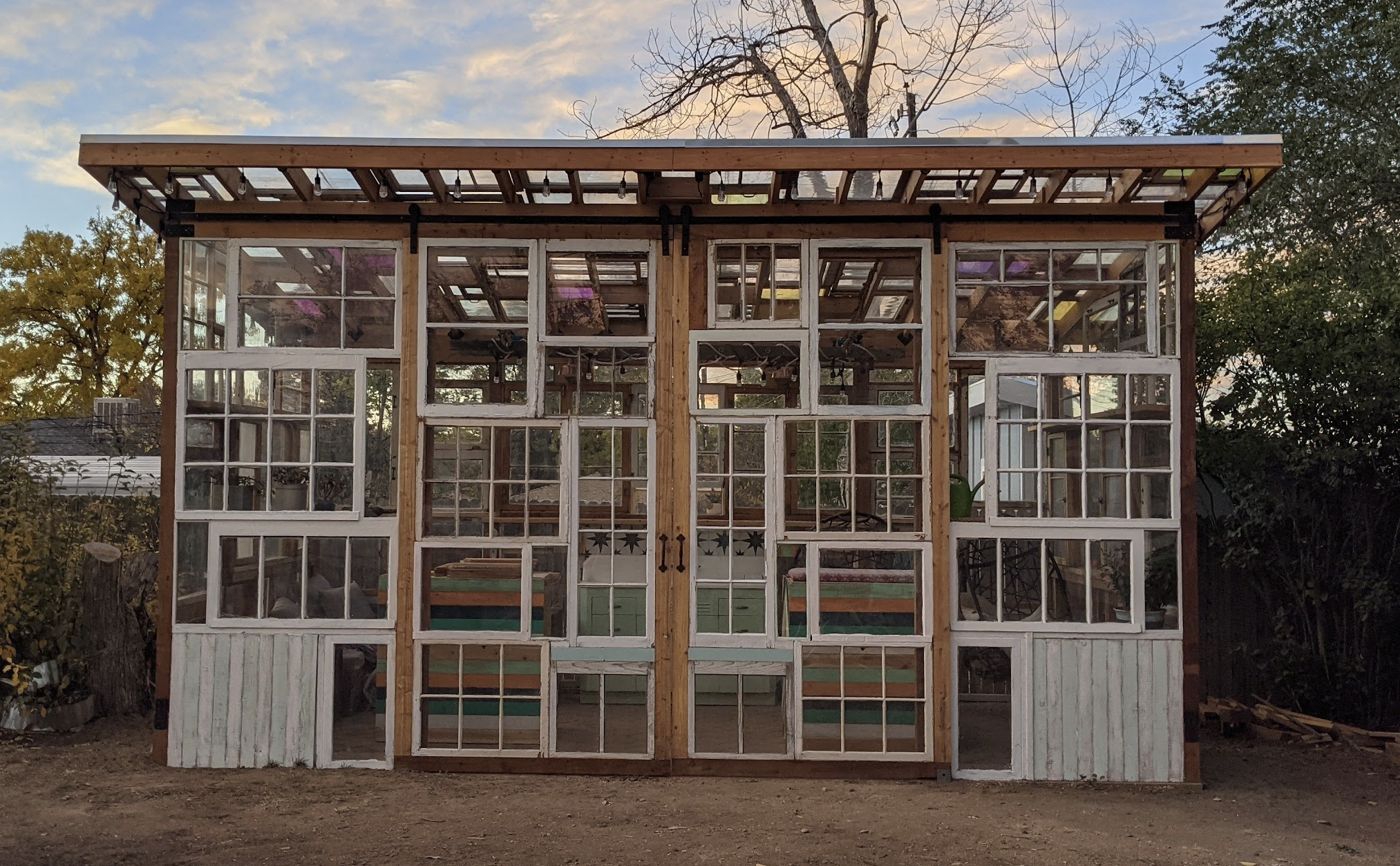 A small, partly constructed greenhouse with a wooden frame and large windows, situated outdoors during sunset.