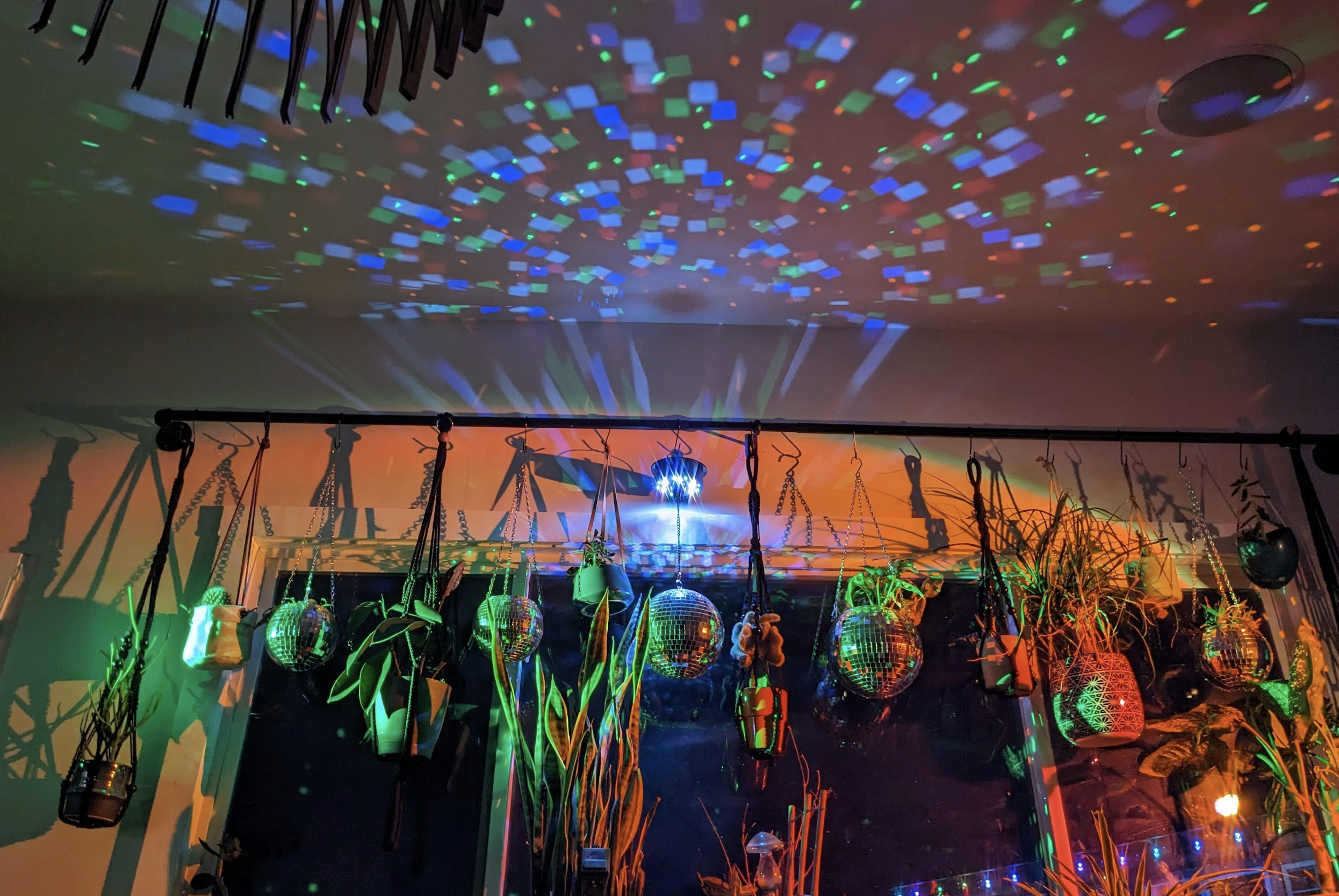 Colorful disco balls and potted plants hanging from a ceiling with multicolored lights projecting patterns onto the ceiling and walls.